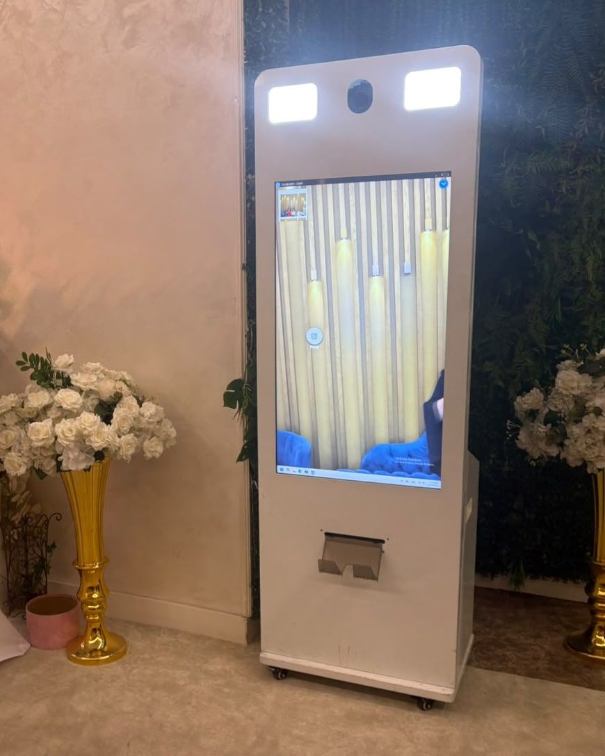 Jovana Photobooth na proslavi: beli photobooth sa ekranom, svetlima i štampačem, između belog cveća u zlatnim vazama